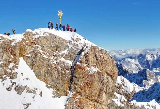 Turyści stoją na skalistym szczycie przy krzyżu z widokiem na ośnieżone Tatry.
